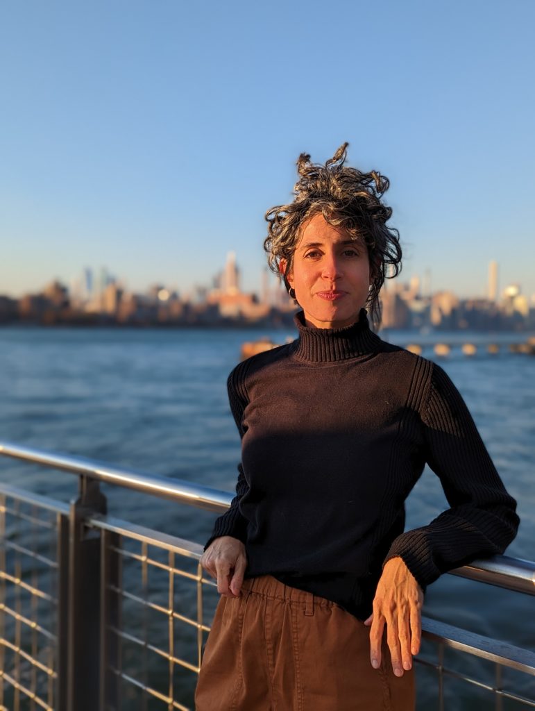 Woman with curly gray curls blowing in the wind wearing a black turtleneck seater and tan pants, leaning with her back to a railing and staring directly at the camera with a slight smile.