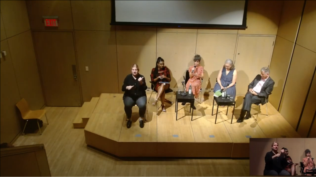 On overview five people sitting on a wooden stage with a screen behind them. One is signing, and one holding the mic.